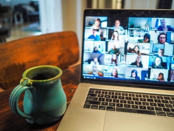 A laptop with people's faces in the usual online meeting squares. A blue pottery mug is next to the keyboard.