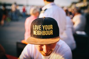 A person, head and shoulders, wearing a white t shirt and a black ball cap reading "love your neighbour".