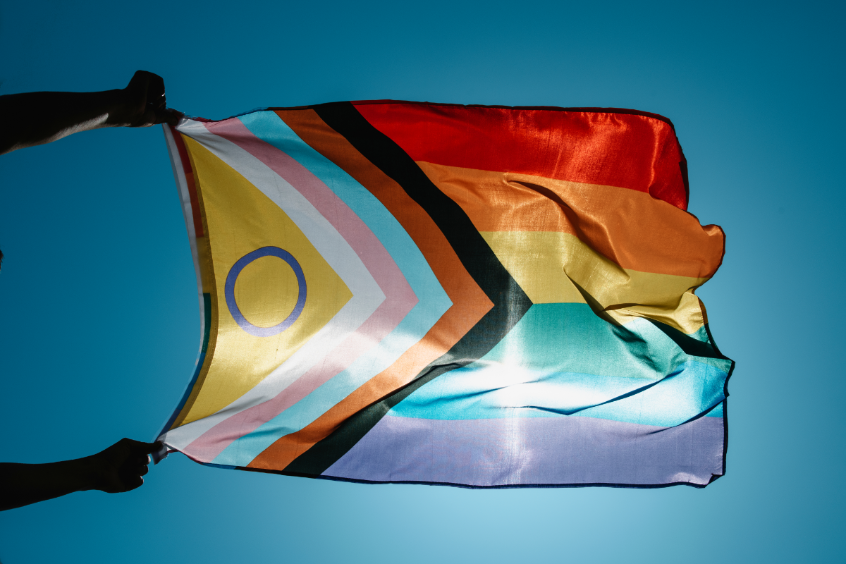 An inclusive Pride flag, with the sun shining through it, with two hands holding it as it blows in the wind.