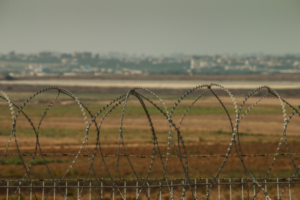 Razor wire in the foreground, a strip of bare brown earth, then a distant line of buildings.