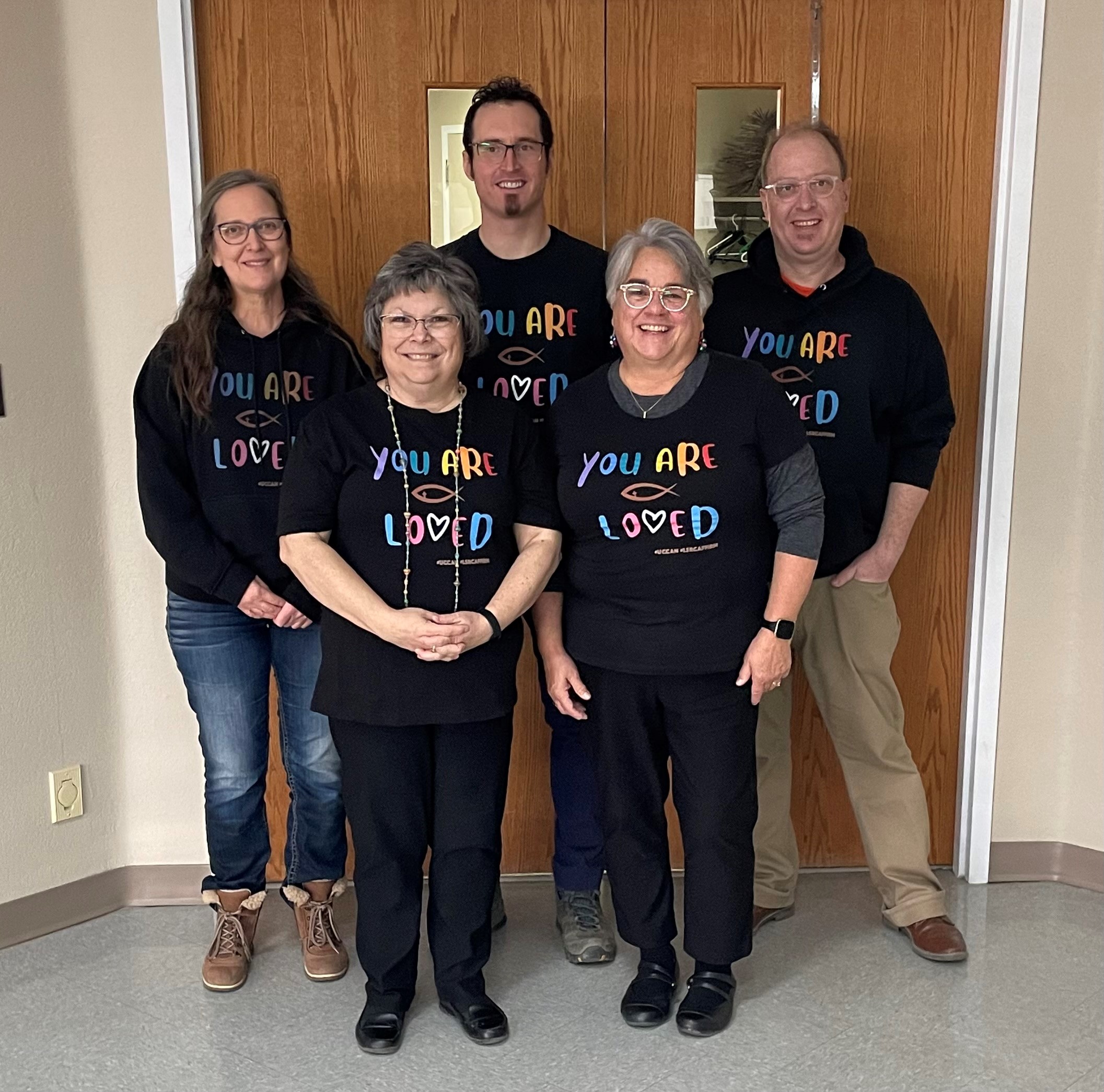 A group of 5 people wearing black t shirts with rainbow lettering reading "You are loved".
