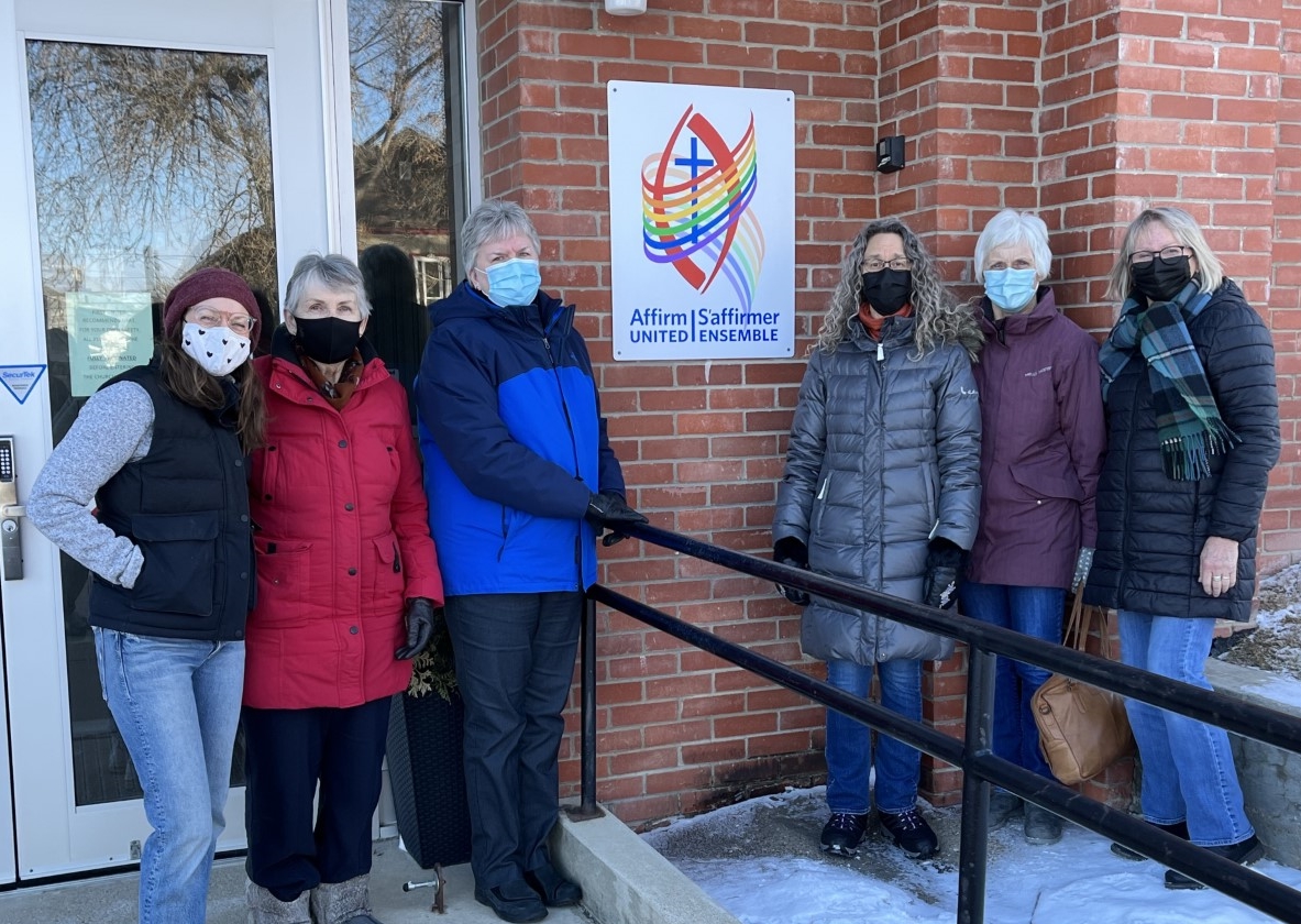 A group of people, bundled against the cold and wearing surgical masks, stand on the church steps next to a large Affirm United logo.