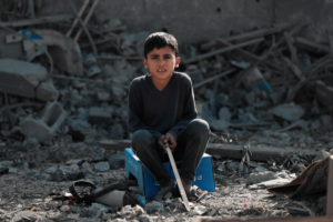 A dark haired child wearing jeans sits on a box surrounded by grey rubble, Gaza.