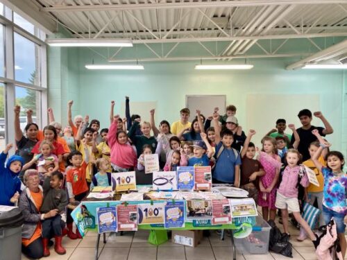 About 30 people gathered around a table in a hall, their hands in the air accompanied by smiles. 